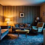 A cozy, retro living room with mismatched vintage furniture, patterned wallpaper, and warm, muted colors. A record player sits on a side table, casting a soft glow